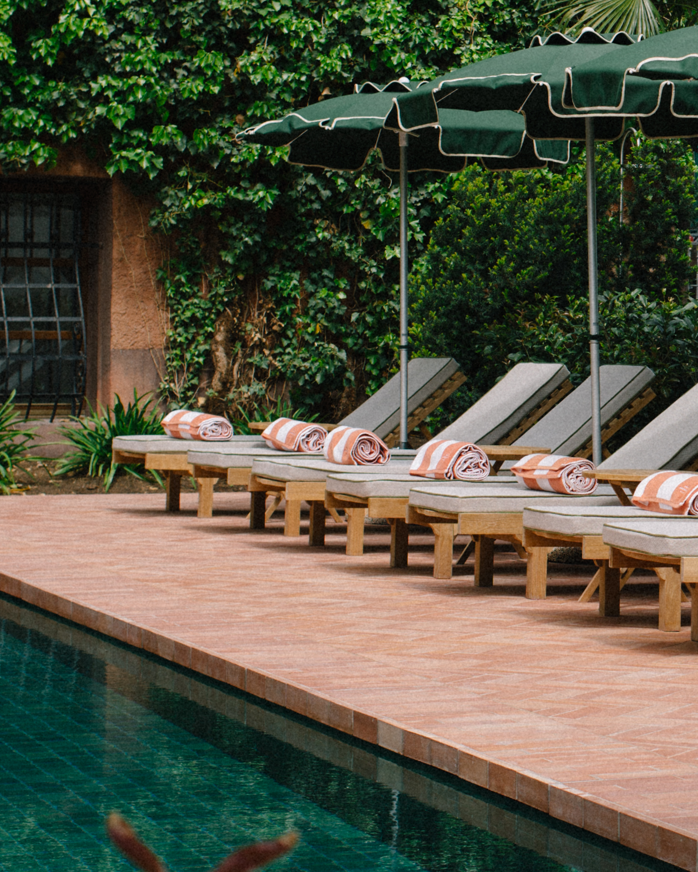 pool loungers with rolled up peach and white striped towels laying on them below green vintage style umbrellas by a pool with pink tiled ground surrounded by green foliage