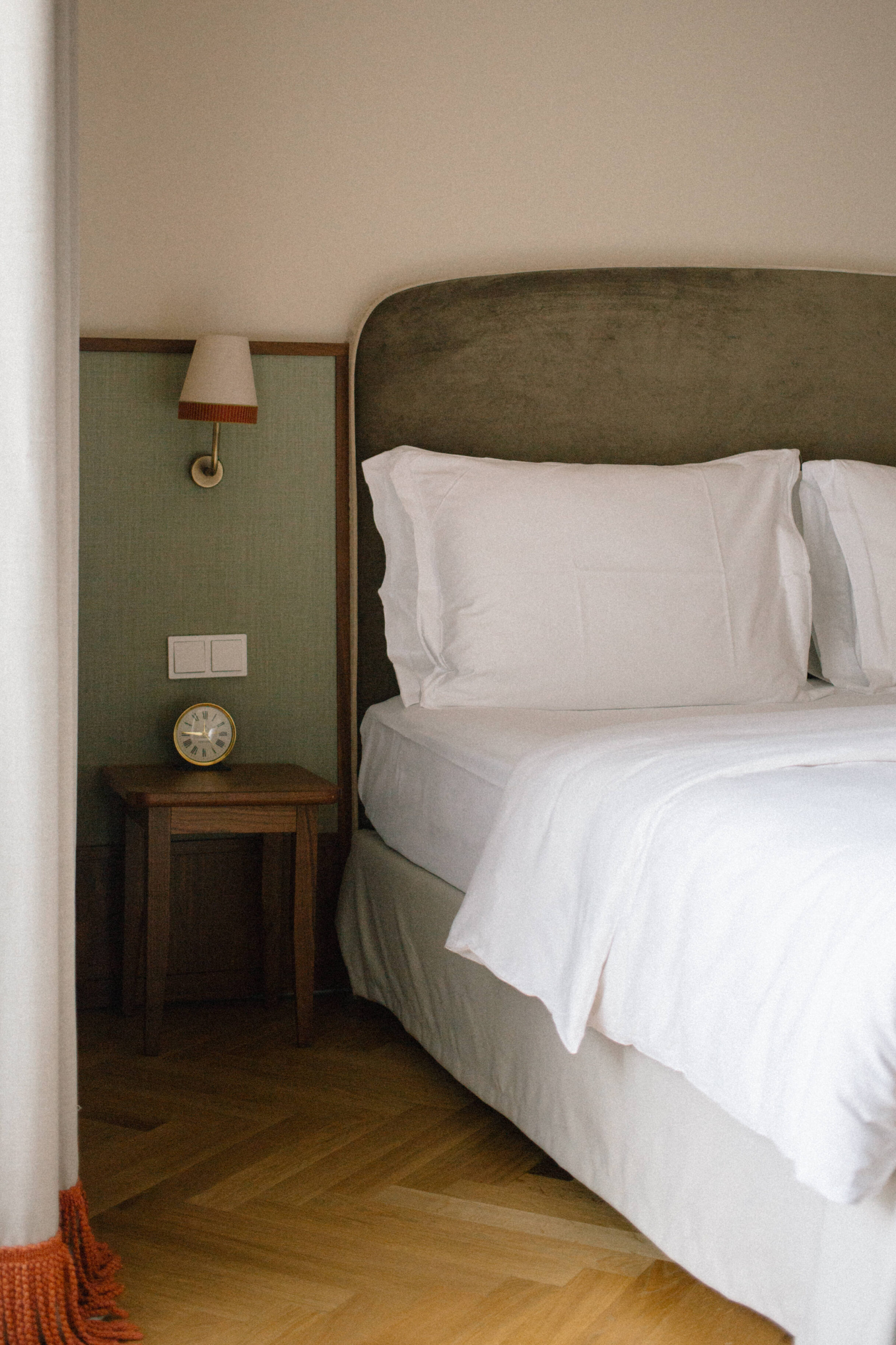 image of a hotel room with a small lamp sconce and green velvet headboard with white sheets and pillow. on the side table there is a vintage clock and light switch