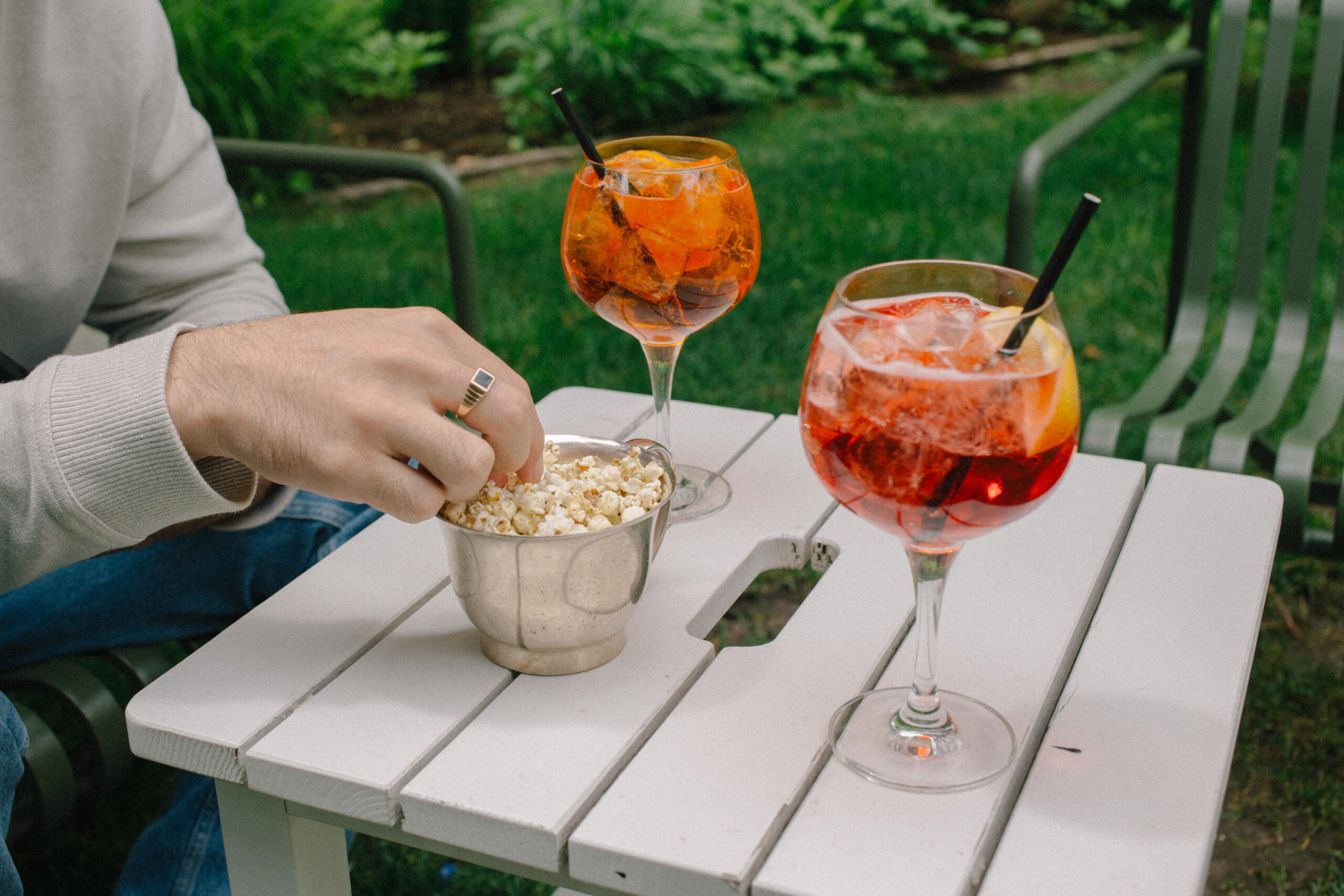 hand of a person grabbing popcorn from a tin bowl on top of a white table with an aperol and campari spritz, the table surrounded by grass and greenery