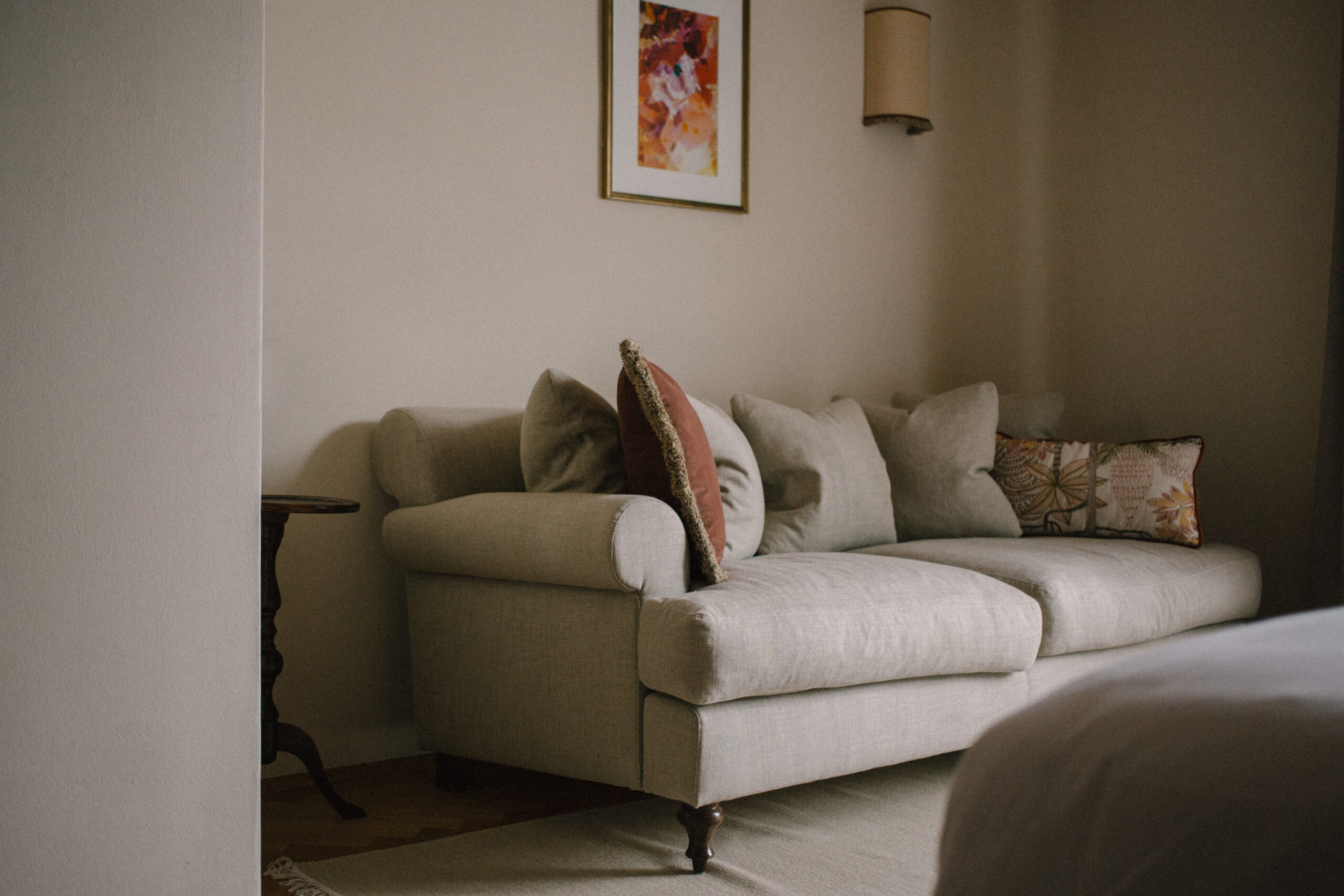 a light grey colored couch in a hotel room with brown accent pillows and wood detailing. on the wall is a framed art piece and canvas wall sconce